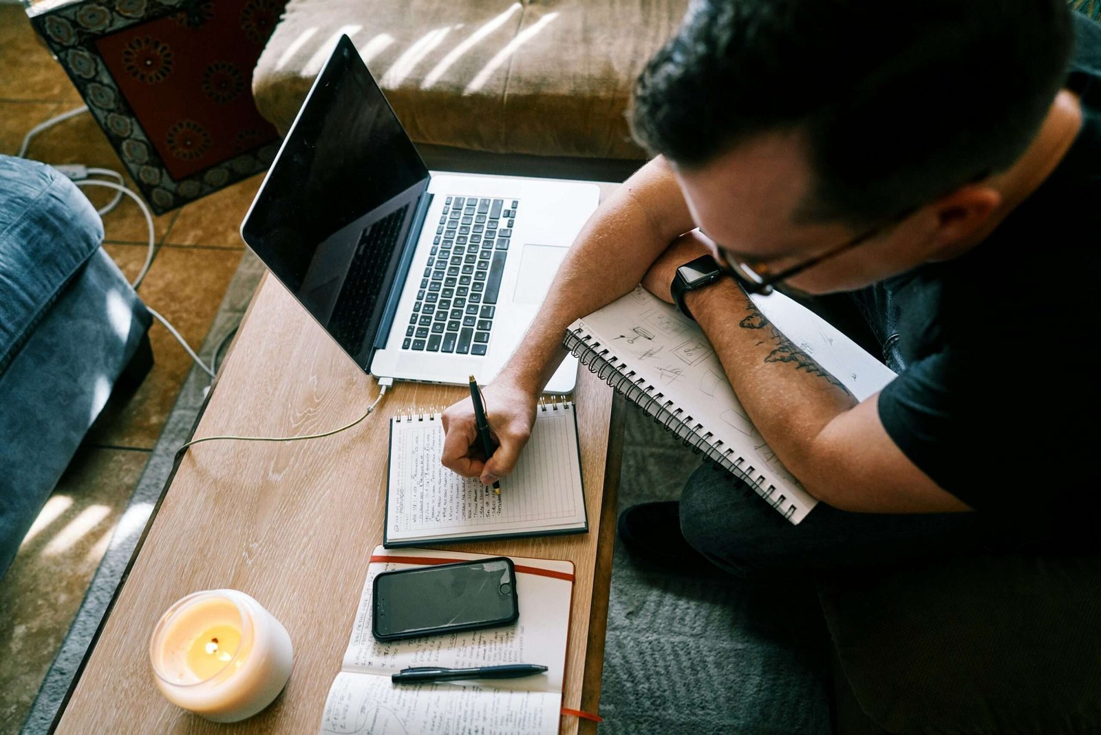 Man studying with laptop, notebook, and smartphone on a wooden table, taking notes.